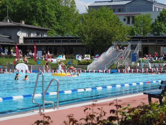 Schwimmerbecken mit rot-weißen-Fähnchen dekoriert. Im Hintergrund sieht man die Freibad-Rutsche und Personen, die im Wasser sind oder auf der Liegewiese entspannen.