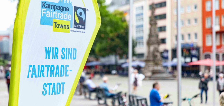 Fairtrade-Banner links, im Hintergrund befinden sich verschwommen Fußgänger, Radfahrer und ein Turm.