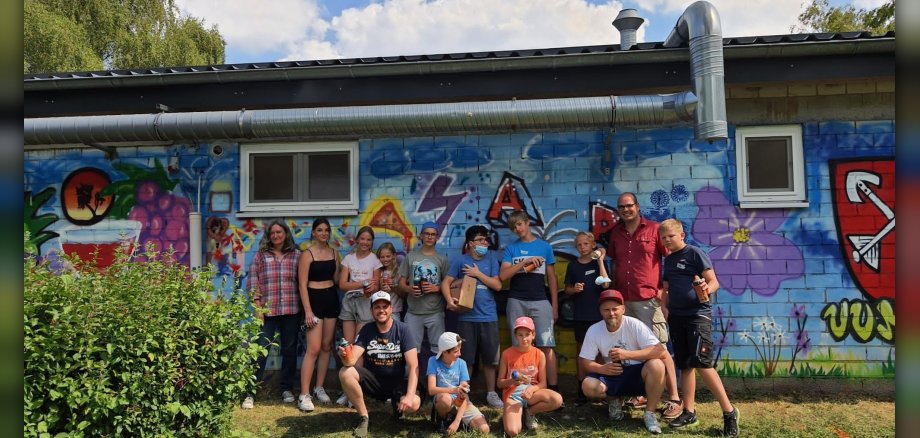 Gruppenfoto der teilnehmenden Kinder und Jugendlichen sowie Organisatoren vor der fertig besprühten Wand.