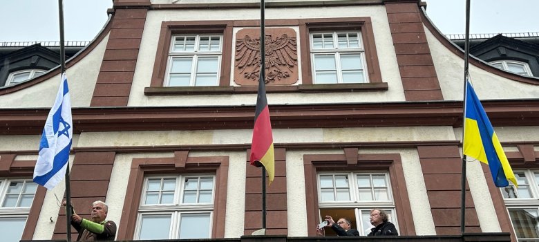 Drei Personen stehen auf dem Balkon des Eltviller Rathauses. Eine Person hisst eine Fahne Israels. Insgesamt sind 3 Flaggen zu sehen, alle davon auf Halbmast.