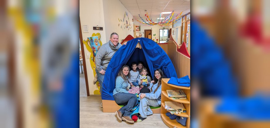 Drei Kinder und drei Frauen in und vor einem Zelt in einem Kindergarten