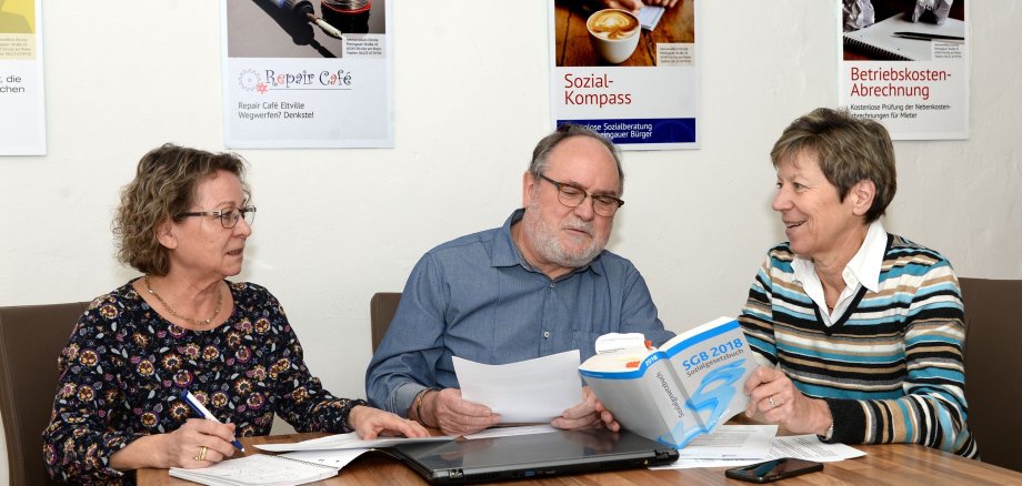 Ein Mann und zwei Frauen sitzen an einem Tisch und betrachten verschiedene Unterlagen. Eine Frau hält das Sozialgesetzbuch in der Hand.